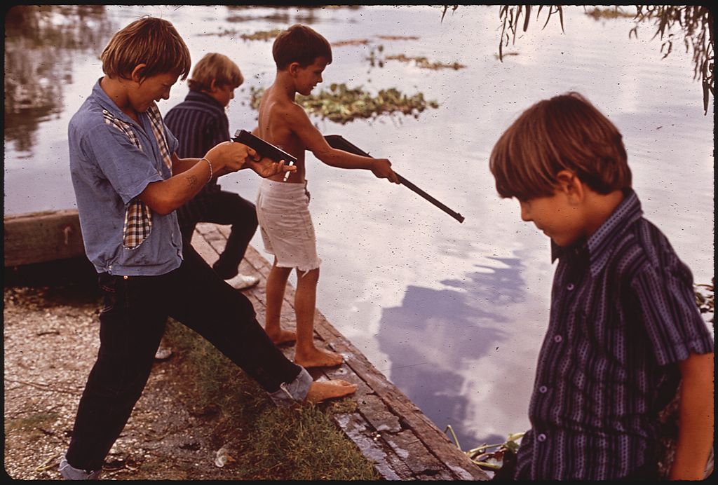 A vintage snapshot of four boys playing with toy guns next to a lake.
