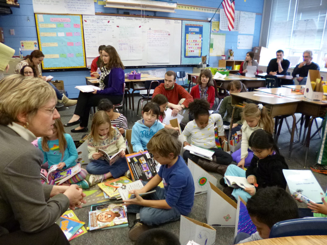 Photograph of a classroom with a teacher in the foreground and children gathered sititing on the floor