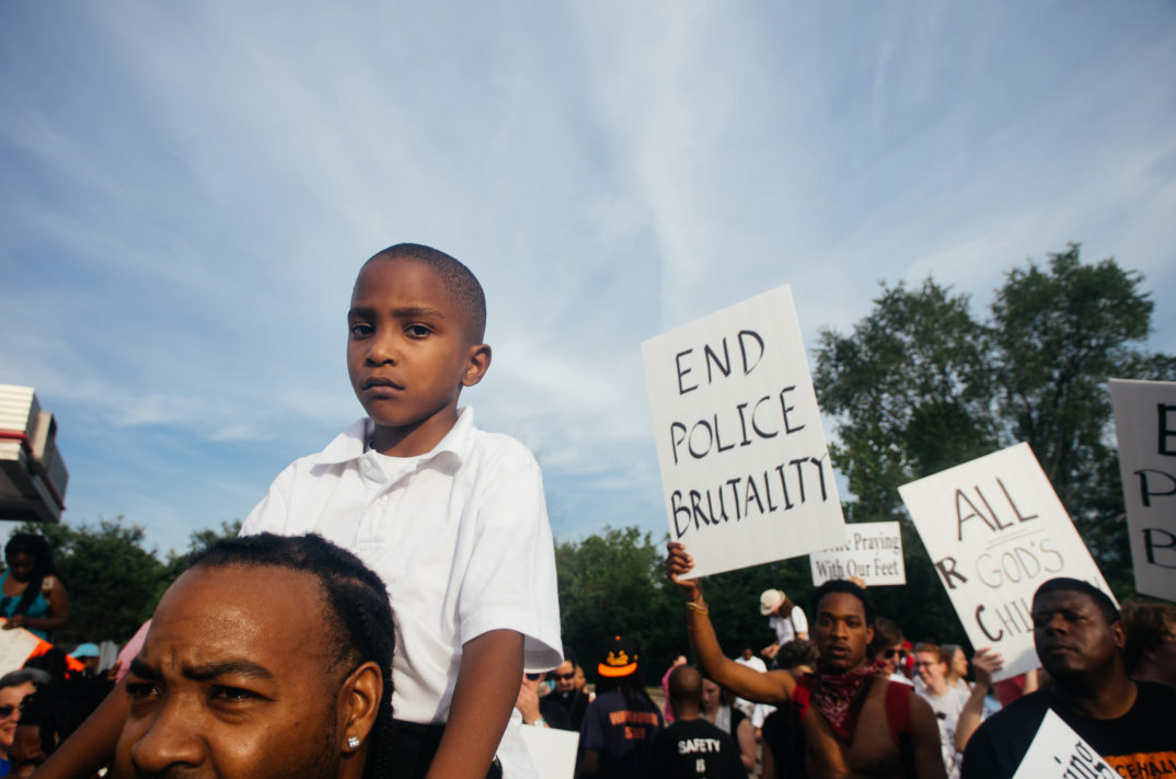 Photograph of protest with boy in foreground, a sign in the background saying "end police brutality"