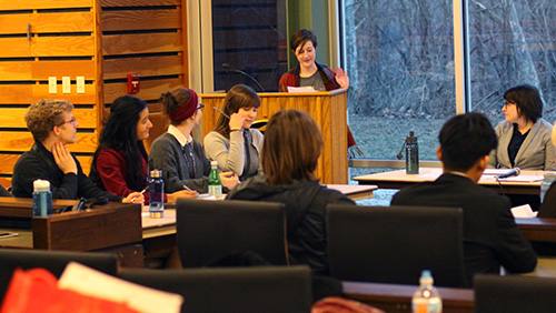 A woman behind a podium addresses a group of students and adults seated at tables