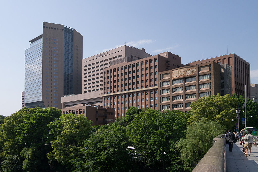 Slightly angled view of the exterior of a large complex of buildings known as the Tokyo Medical and Dental University with a stand of trees in front