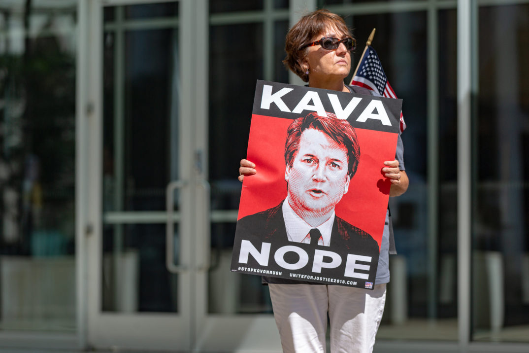 photograph of a woman holding a sign with the slogan "kava-nope" and a picture of Brett Kavanaugh