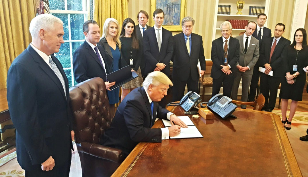 PHotograph of Trump at desk in Oval Office surrounded by people