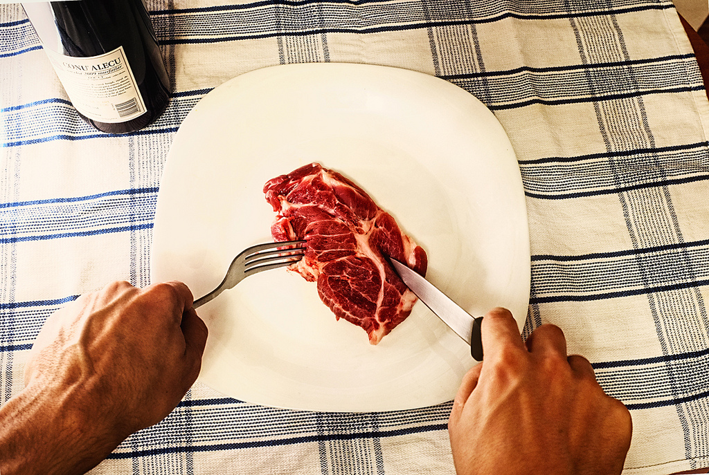 Photograph of a person's hands holding a knife and fork with a piece of raw meat on a plate beneath the utensils