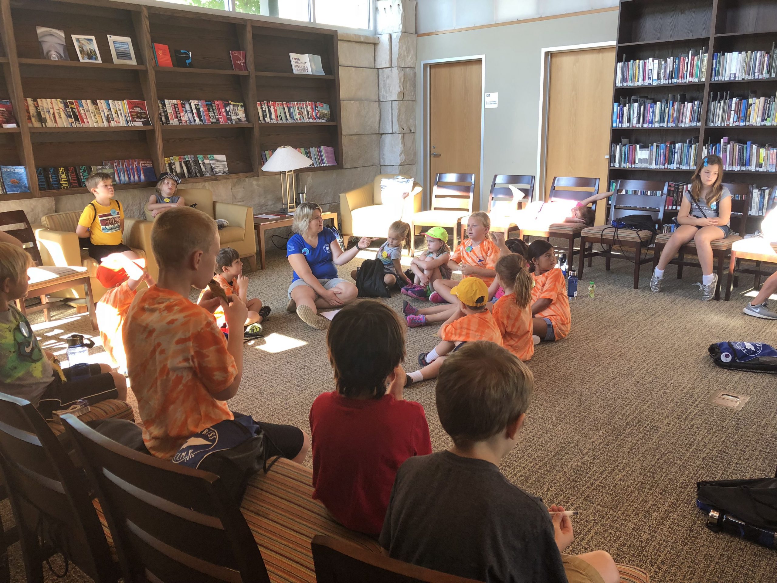 A large group of children, many of whom wear tie-dyed orange shirts gather in a semicircle in a library setting around a teacher