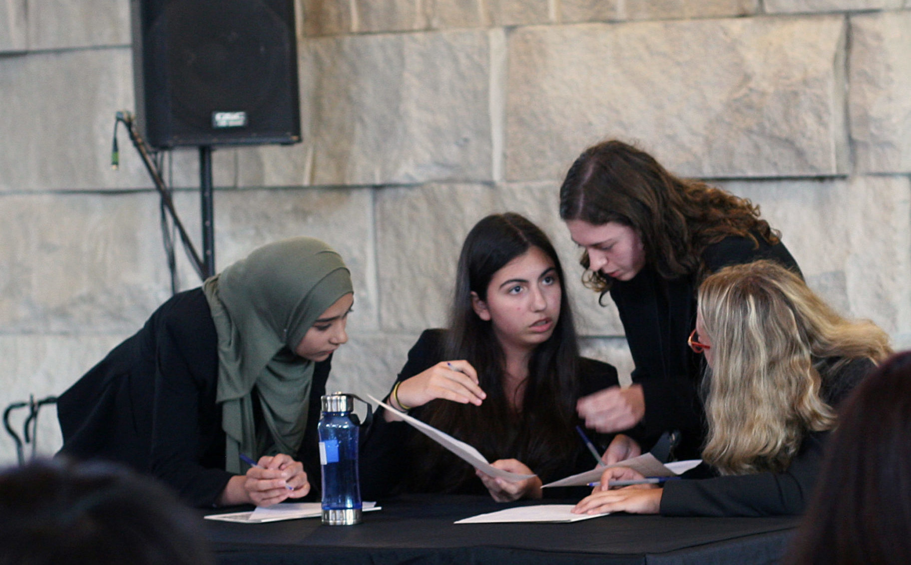 A group of high school students lean into one another around a table to discuss their ethics bowl case.