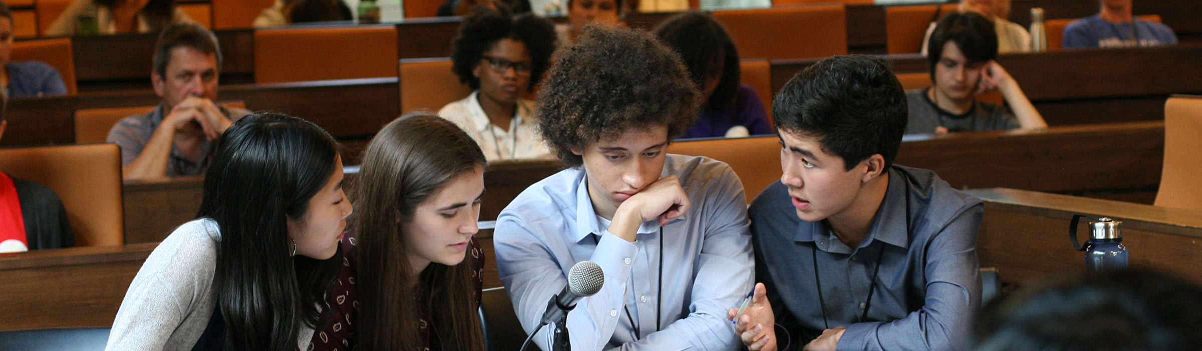 A group of high school students lean into one another around a table to discuss their ethics bowl case.