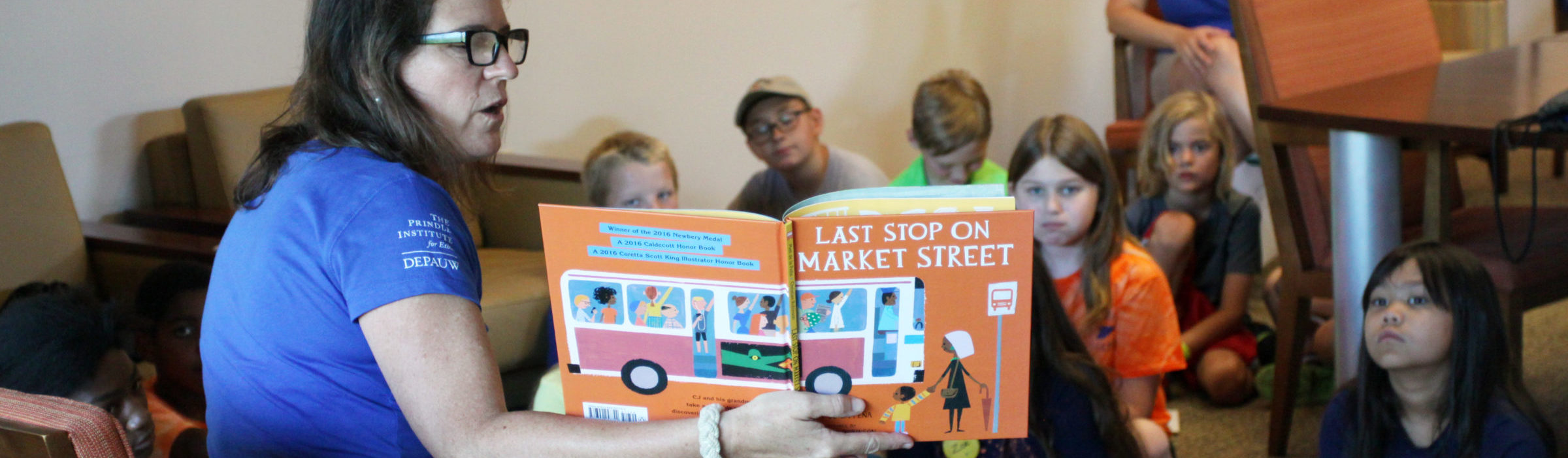 A teacher on a chair in a blue camp shirt, reads Last Stop On Market to a group of 7 students sitting on the floor, looking at the book. Another teacher, in a matching blue shirt, looks on from behind the students.