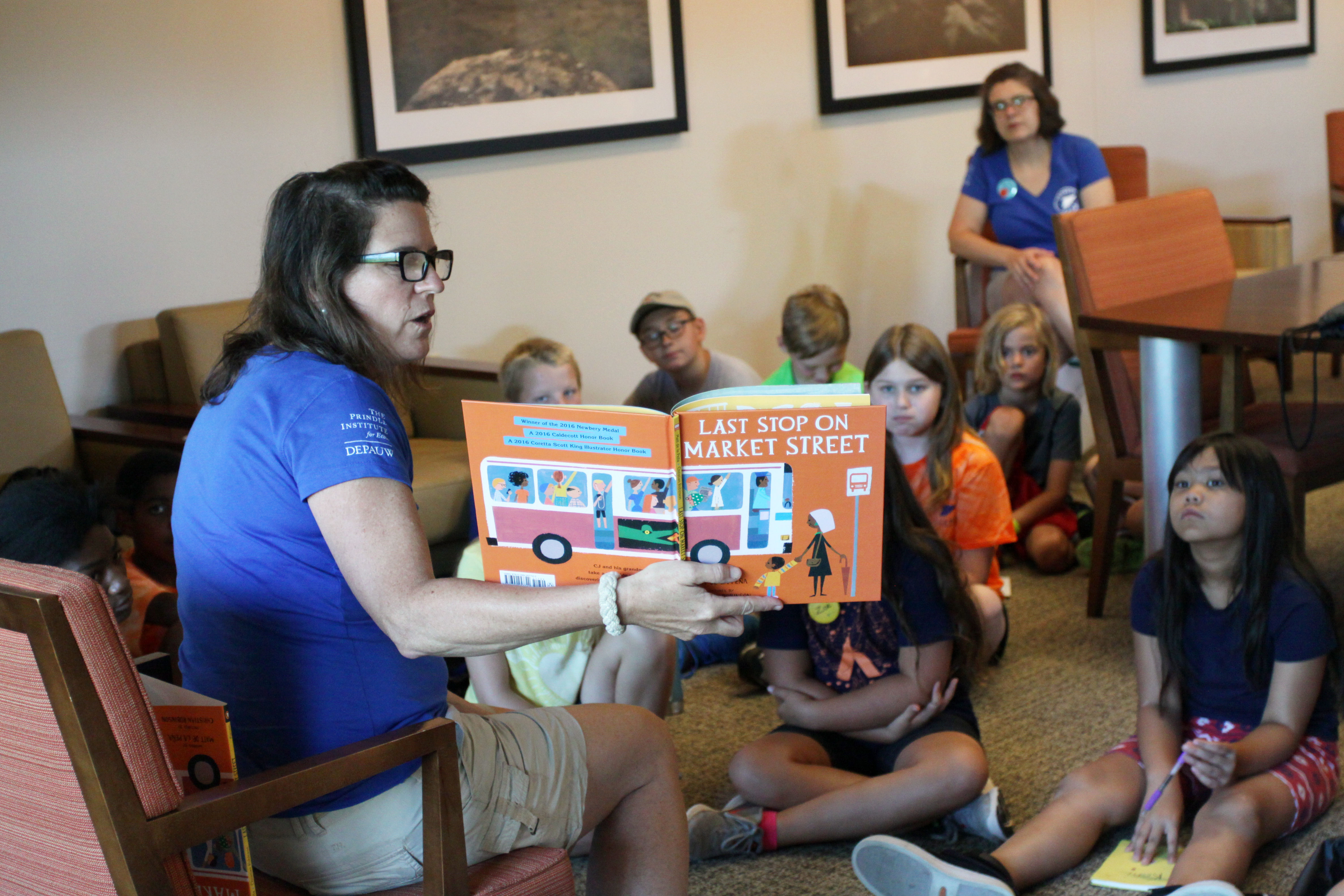 A teacher on a chair in a blue camp shirt, reads Last Stop On Market to a group of 7 students sitting on the floor, looking at the book. Another teacher, in a matching blue shirt, looks on from behind the students.