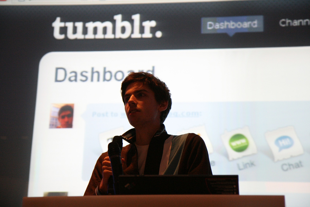 Photo of a man speaking into a microphone, standing in front of a screen displaying a tumblr dashboard