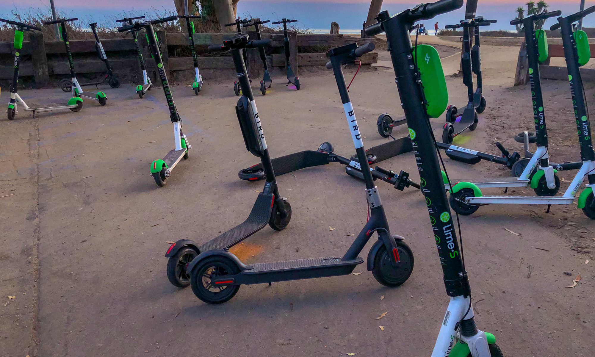 Photograph of several dockless scooters on an area of pavement with a sunset sky in the background