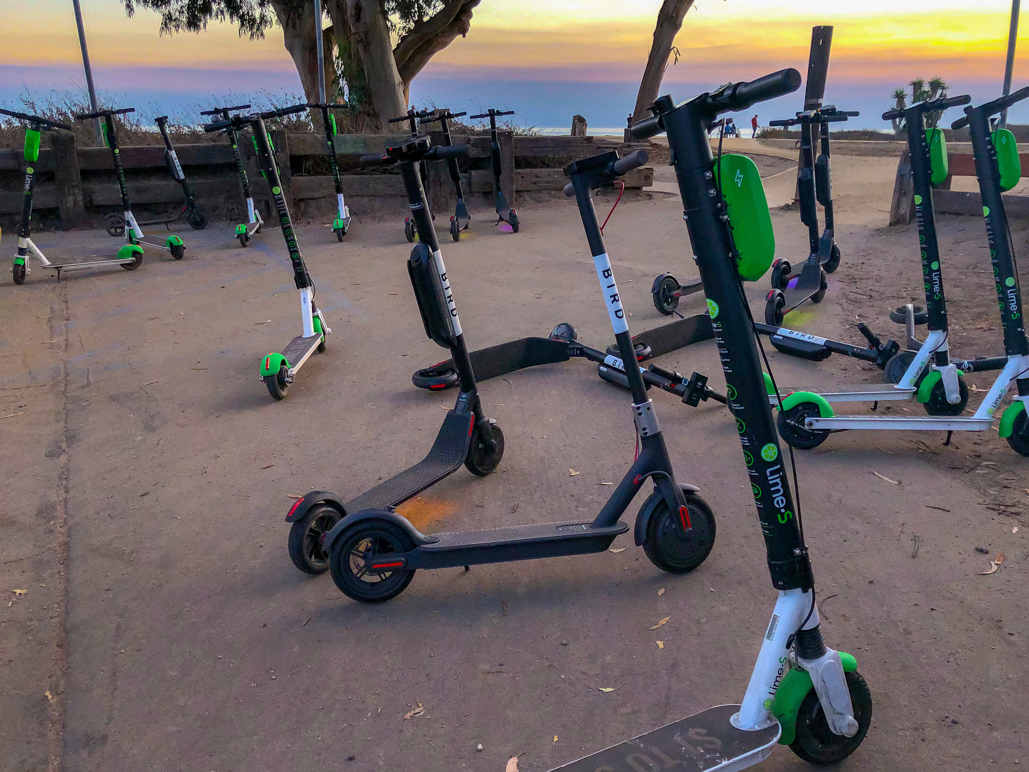 Photograph of several dockless scooters on an area of pavement with a sunset sky in the background