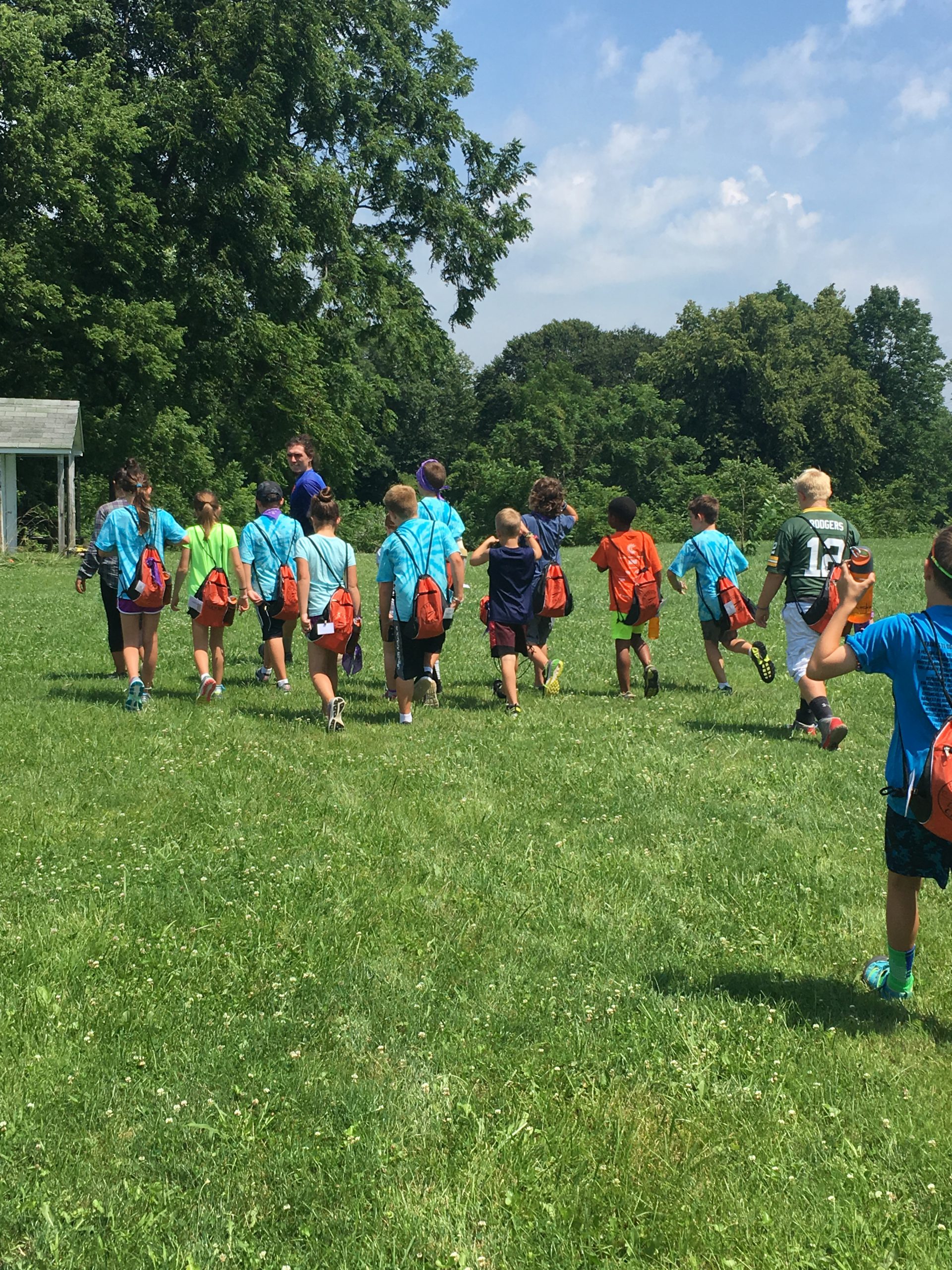 A large group of elementary-school aged children in shorts and short-sleeved shirts walking away from the viewer on a large grassy field.