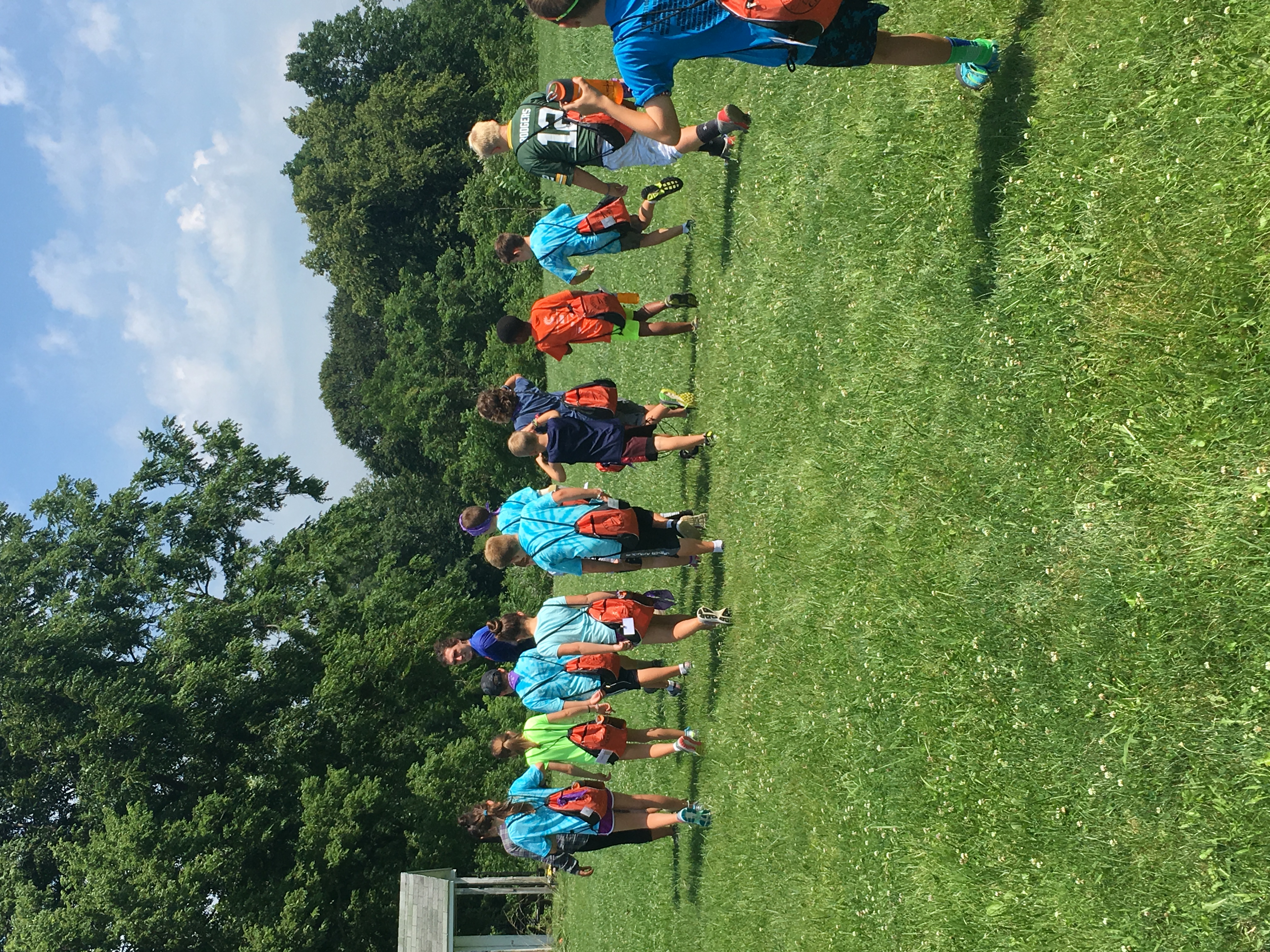 A large group of elementary-school aged children in shorts and short-sleeved shirts walking away from the viewer on a large grassy field.