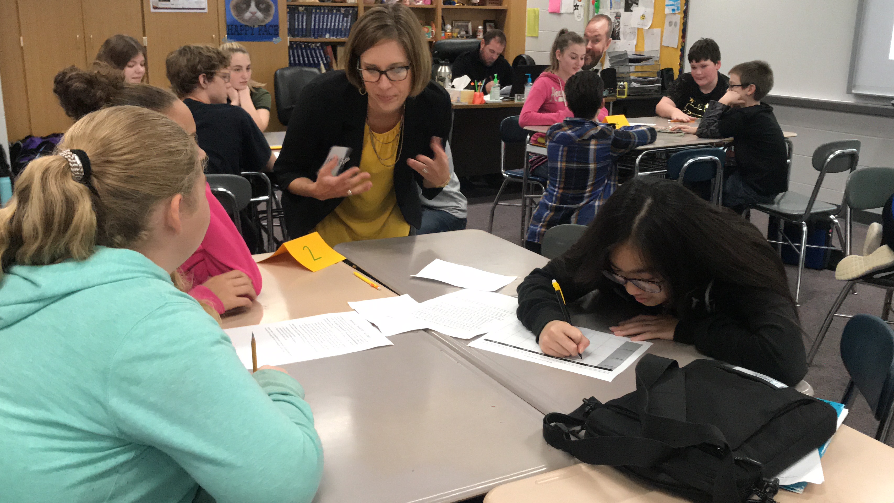 In a crowded classroom, a Prindle staff member leans forward to engage a group of students seated around a cluster of desks