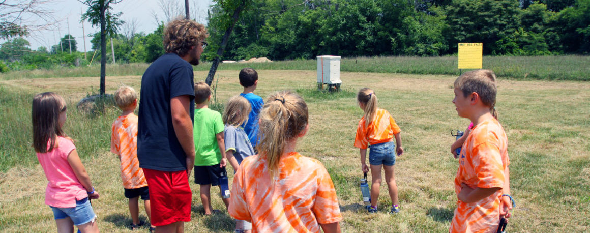 A group of 10 children gather in a circle around a college-age student in a grassy field.