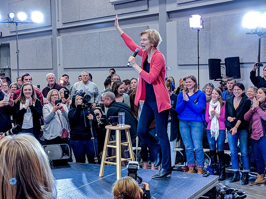 Elizabeth Warren standing on a podium with a stool, speaking to a crowd while holding a microphone