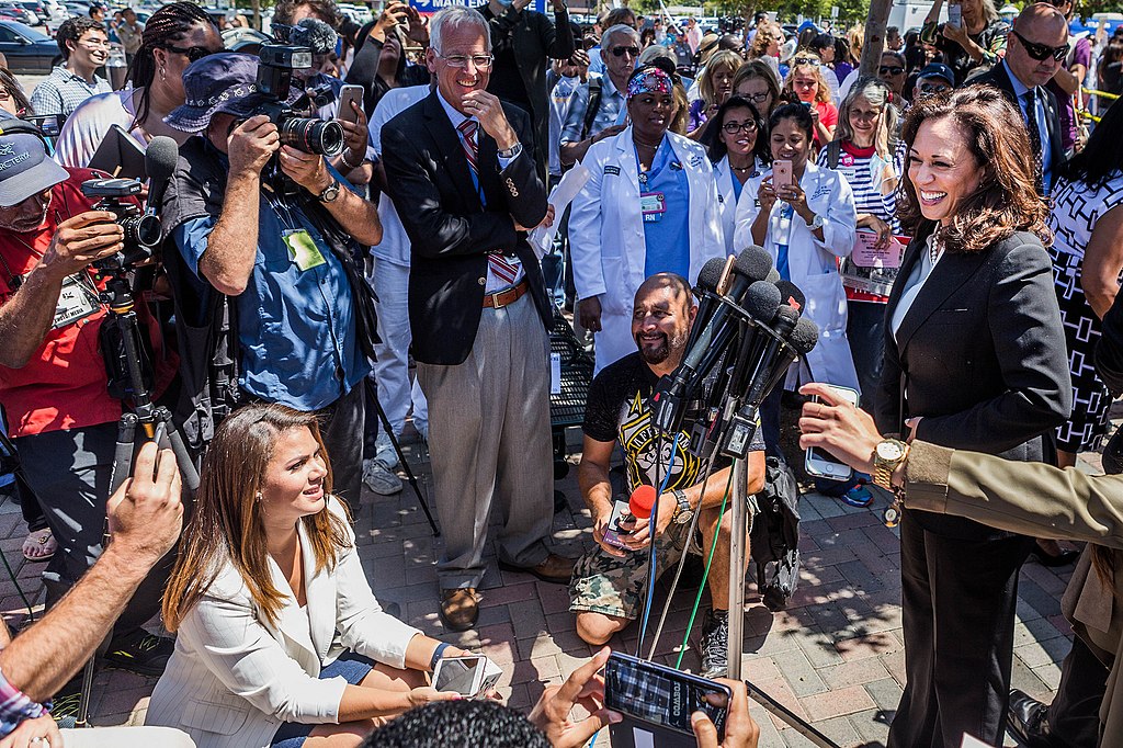 Kamala Harris giving a speech, smiling and speaking into microphones, with people crowded around