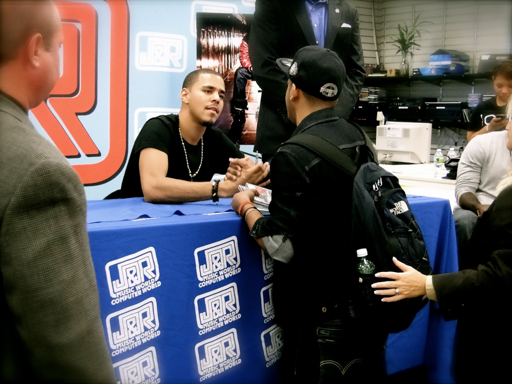 J. Cole sitting at a signing table shaking the hand of a fan