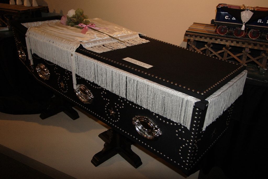 Photograph of a replica of Lincoln's coffin. It is black and draped with white cloth and has a few flowers on top