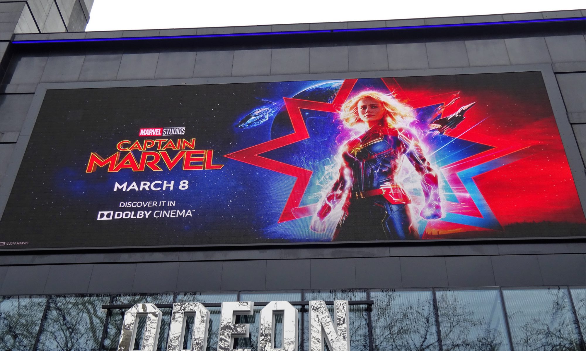 Photograph of a Captain Marvel poster above a movie theatre entrance; the poster shows Brie Larson as Captain Marvel standing with a star and flashing lights behind her