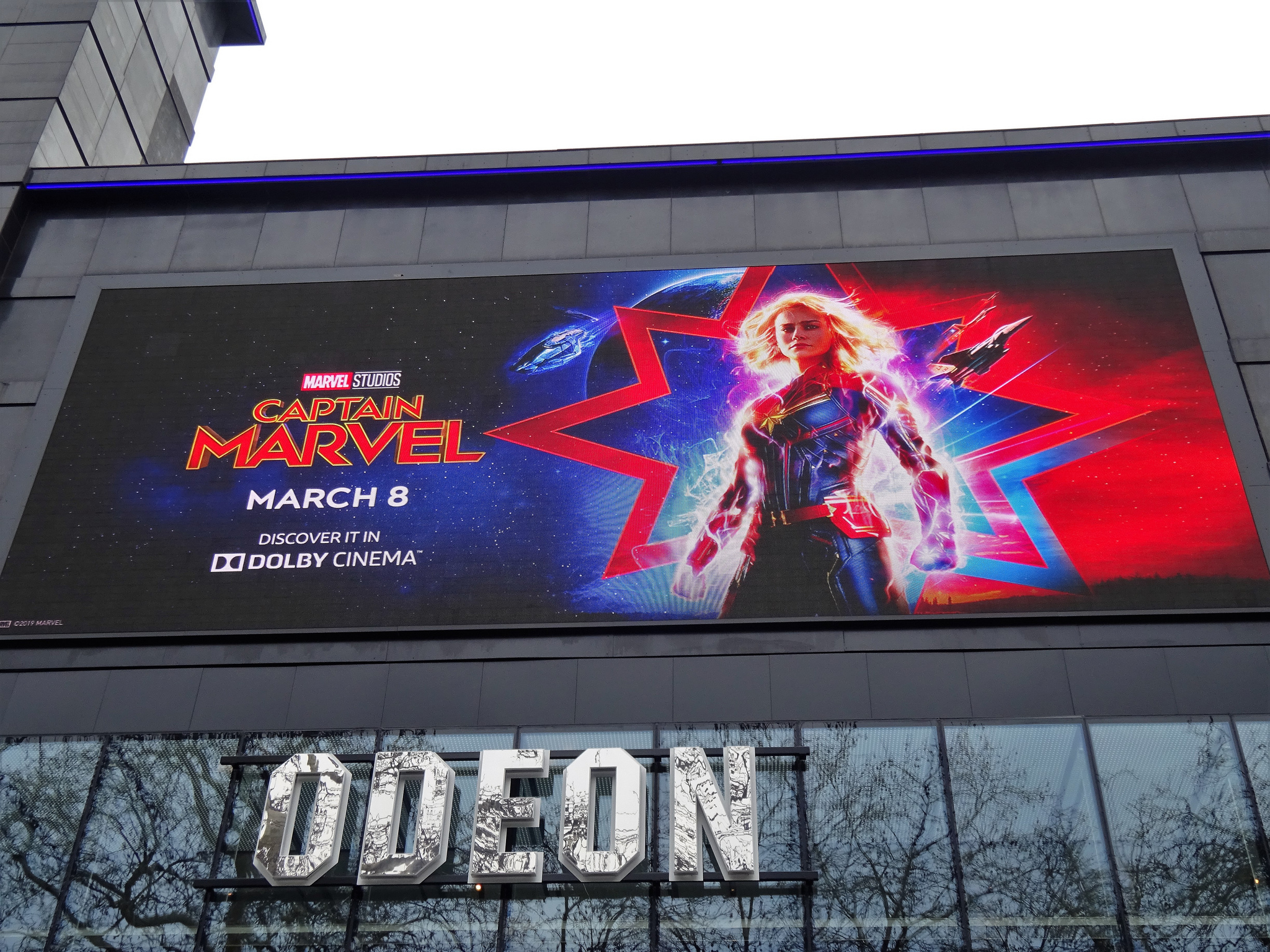 Photograph of a Captain Marvel poster above a movie theatre entrance; the poster shows Brie Larson as Captain Marvel standing with a star and flashing lights behind her