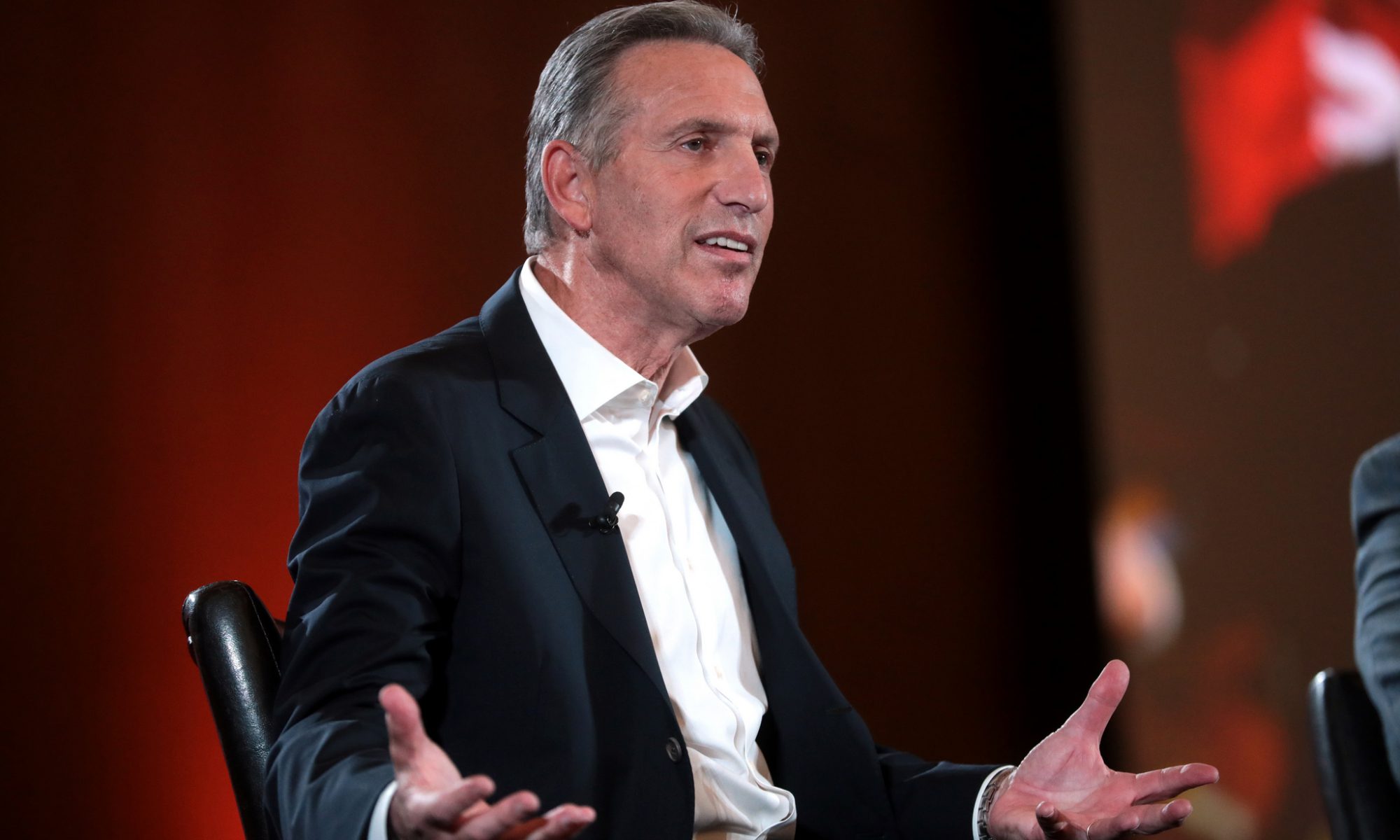 Photograph of former Starbucks CEO sitting on a stage gesturing with his hands spread