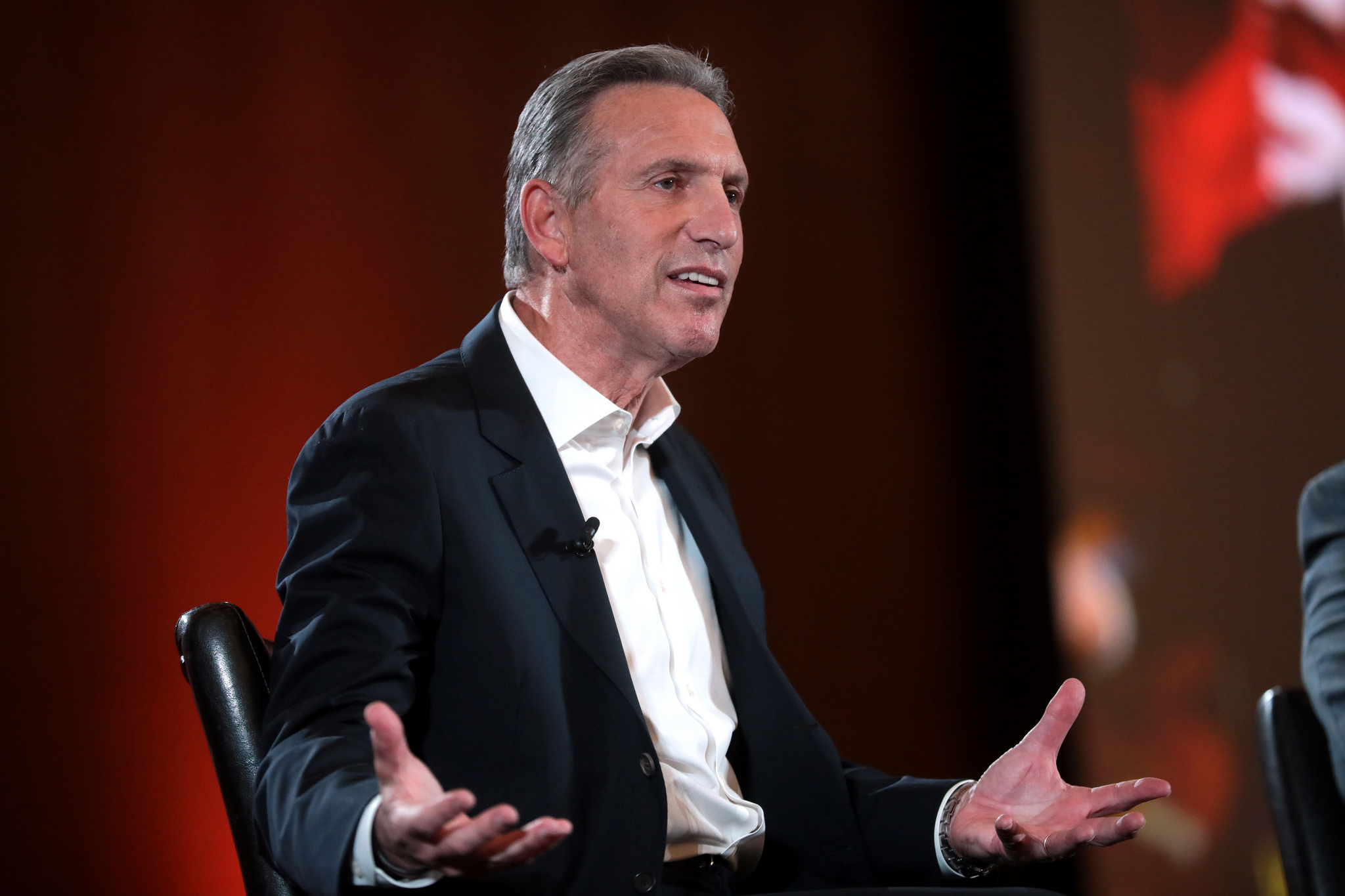 Photograph of former Starbucks CEO sitting on a stage gesturing with his hands spread