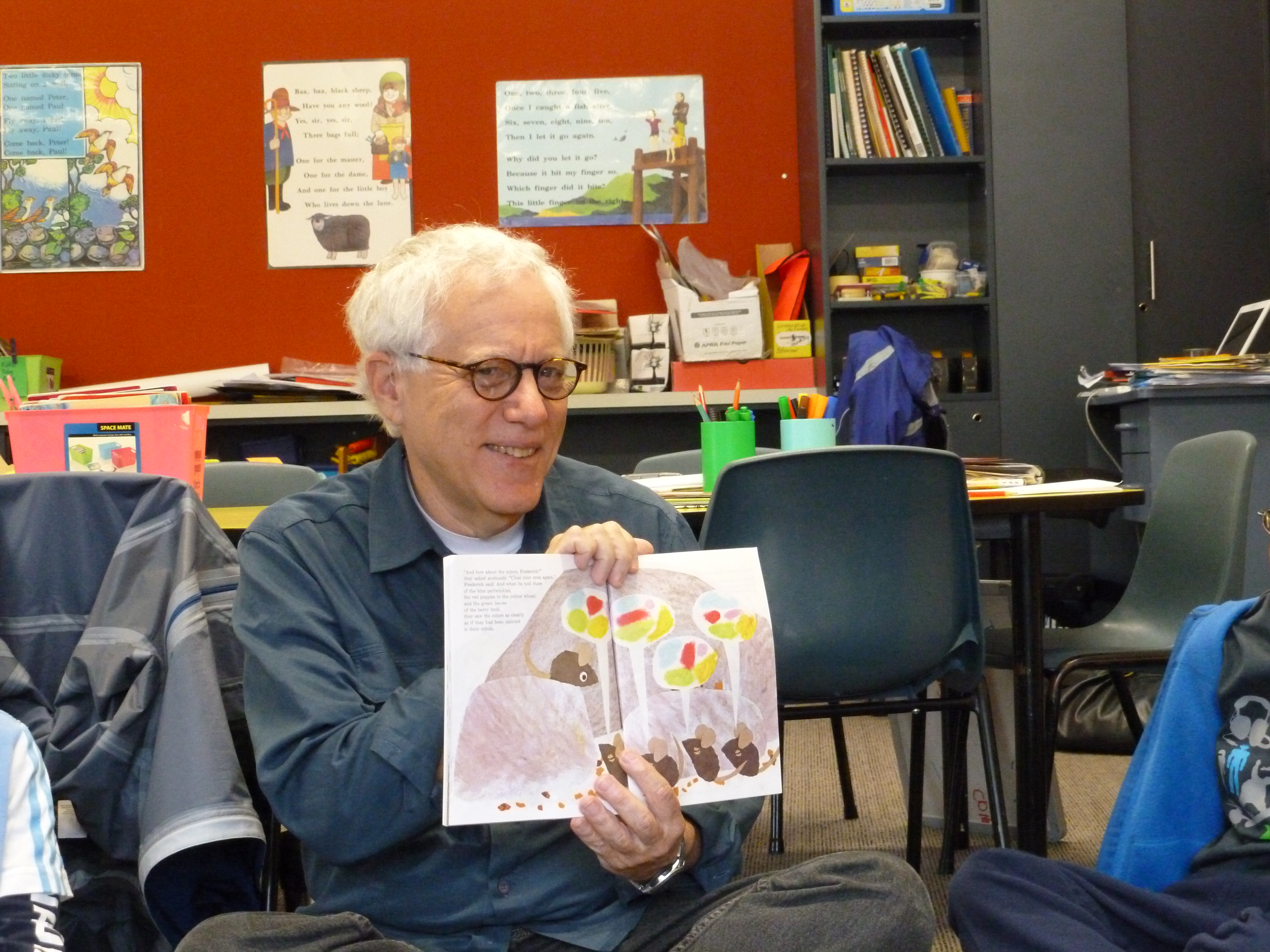A white-haired man with dark glasses smiles as he holds open a children's picture book in a classroom setting.