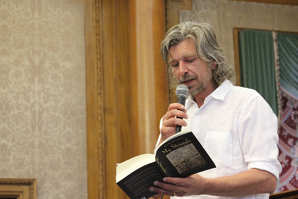 Photograph of author Karl Ove Knausgard standing, holding a microphone, and reading from a book where the title "My Struggle" is visible