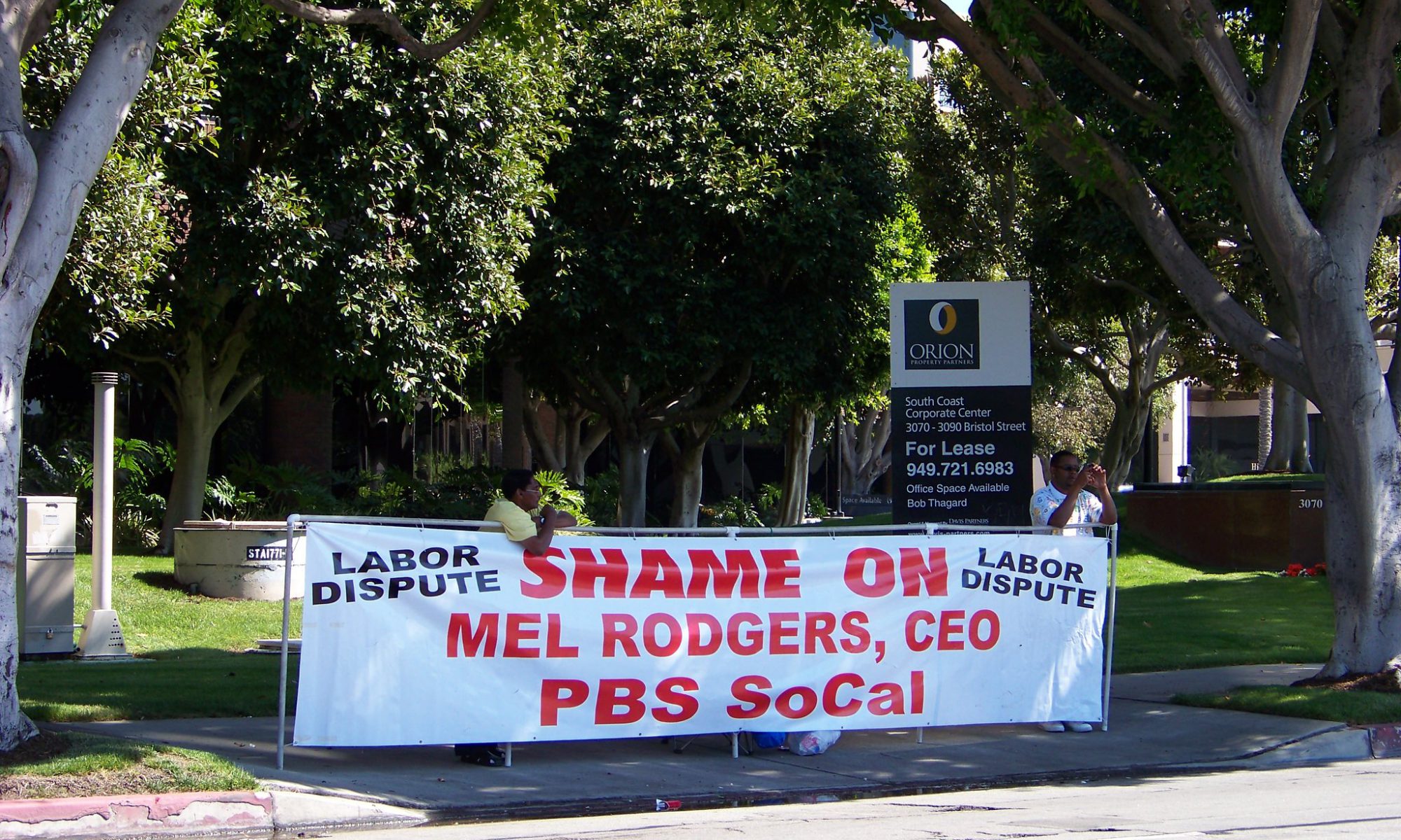 Photo of a person behind a banner that says "Shame on Mel Rogers, CEO, PBS SoCal"