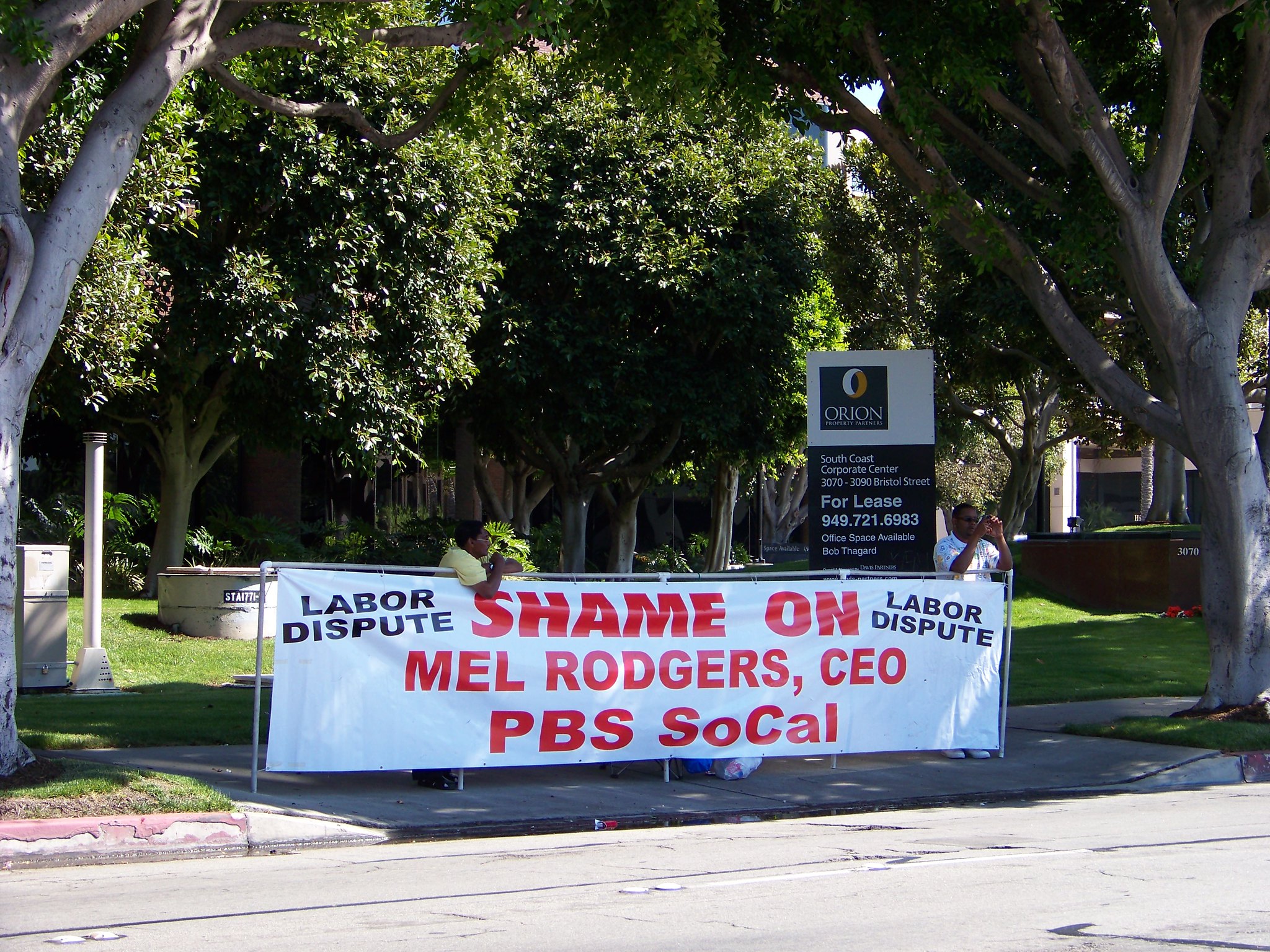 Photo of a person behind a banner that says "Shame on Mel Rogers, CEO, PBS SoCal"
