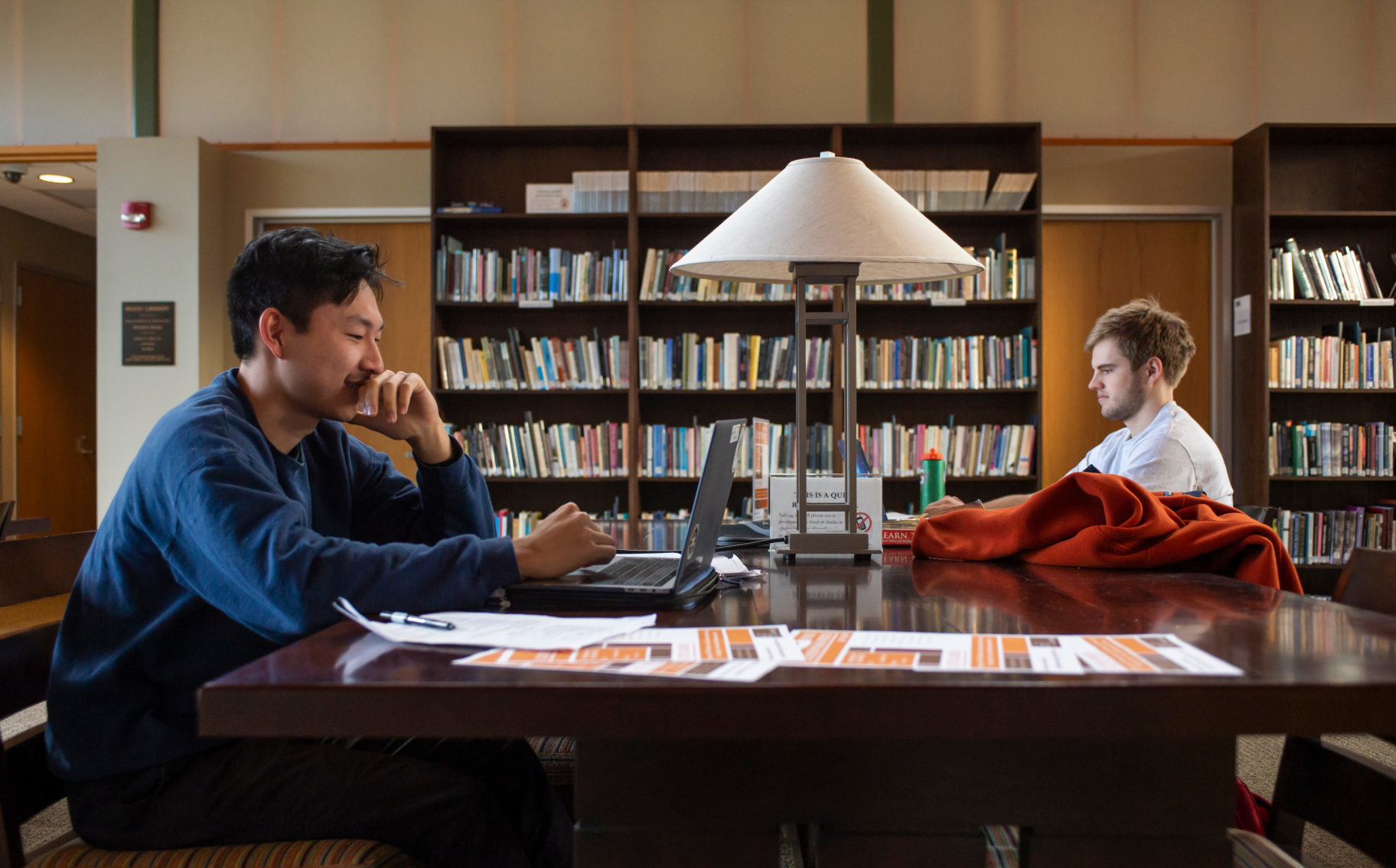 Two students sit across from one another and work on their laptops at a large table in a library.