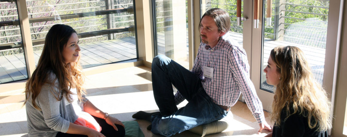 Three people sitting cross-legged on the floor listening intently to one another.