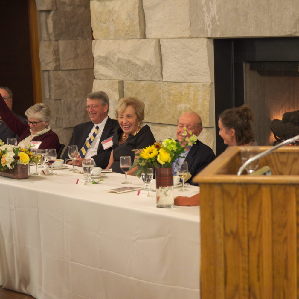 Five people in dress clothes sit behind a long table covered in a white tablecloth. One woman smiles and waves while the others look on with smiles.