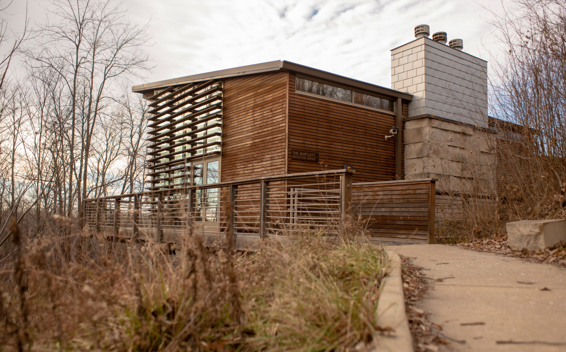 A wooden and glass structure with touches of stone. A sidewalk surrounded by grass and plants leads up to the building.