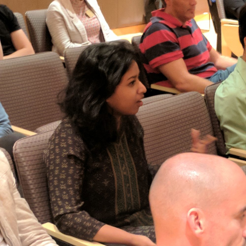 A woman in a crowded auditorium asks a question of the speaker who is off camera