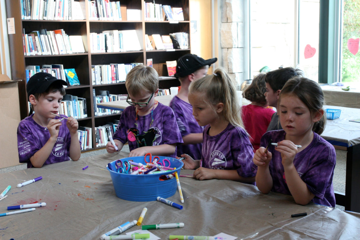 A group of young school children in purple tie-dyed shirts gather around a table and paint small objects.