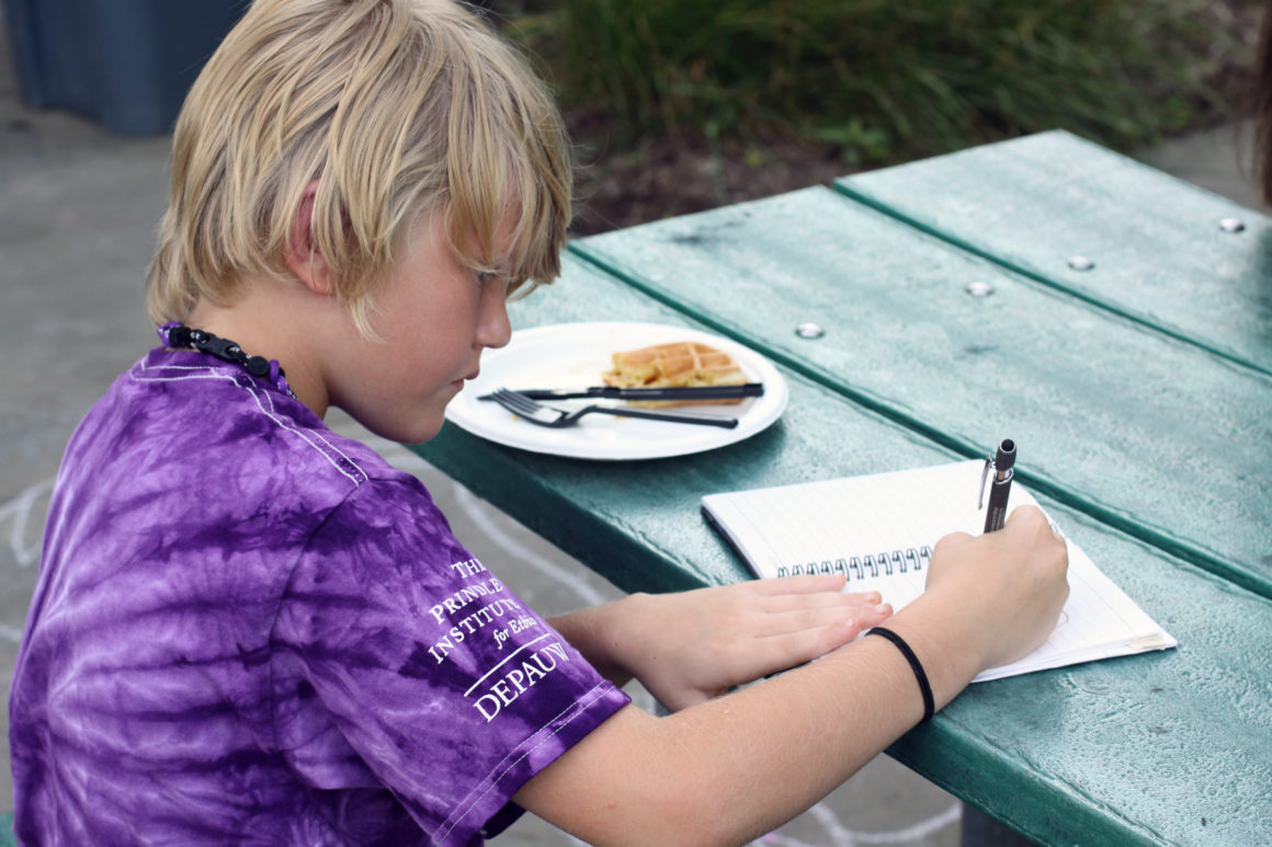 A young child in a purple tie-dye shirt writes in a notebook at a picnic table next to a plate of waffles