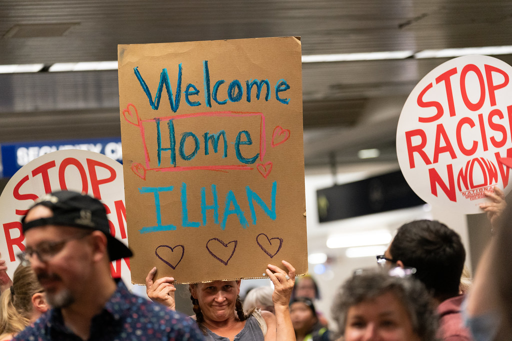 photograph of "Welcome Home Ilhan" sign held by supporter with others gathered at MSP