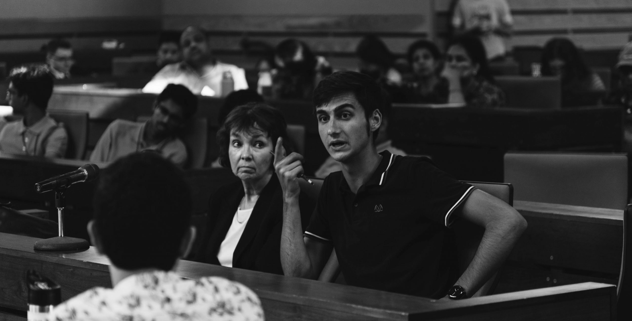 Black and white image of a man speaking while gesturing with his hand. A woman is seating next to him.