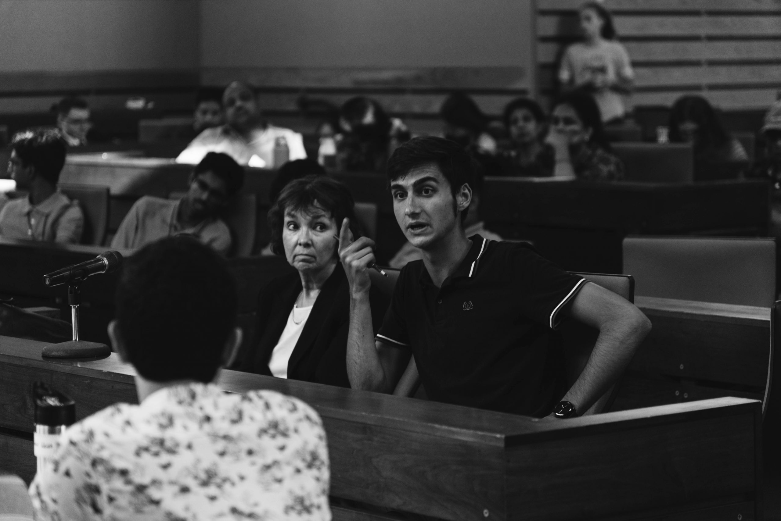 Black and white image of a man speaking while gesturing with his hand. A woman is seating next to him.