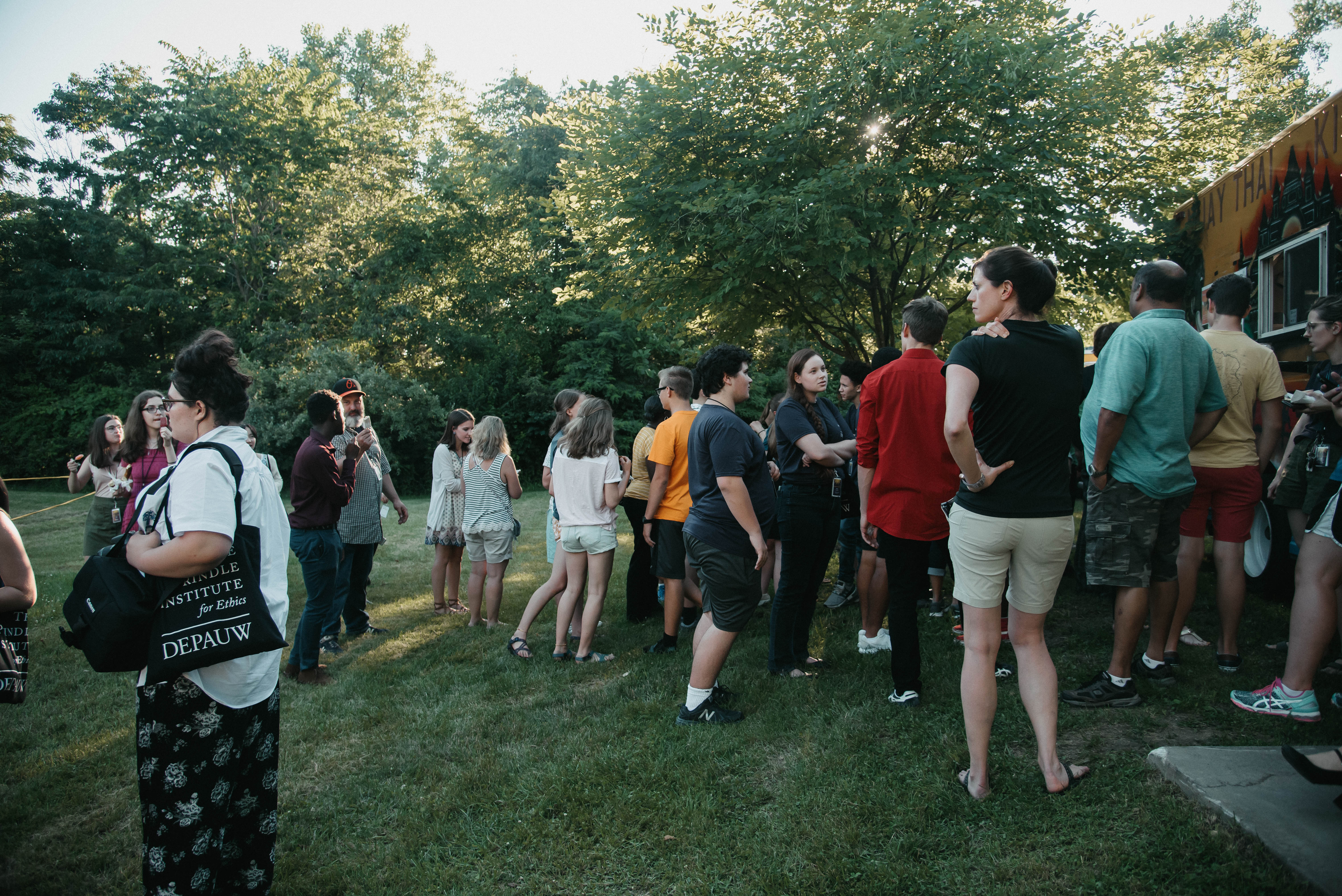 A large group of young students and adults stand in line for a food truck on a grassy field.