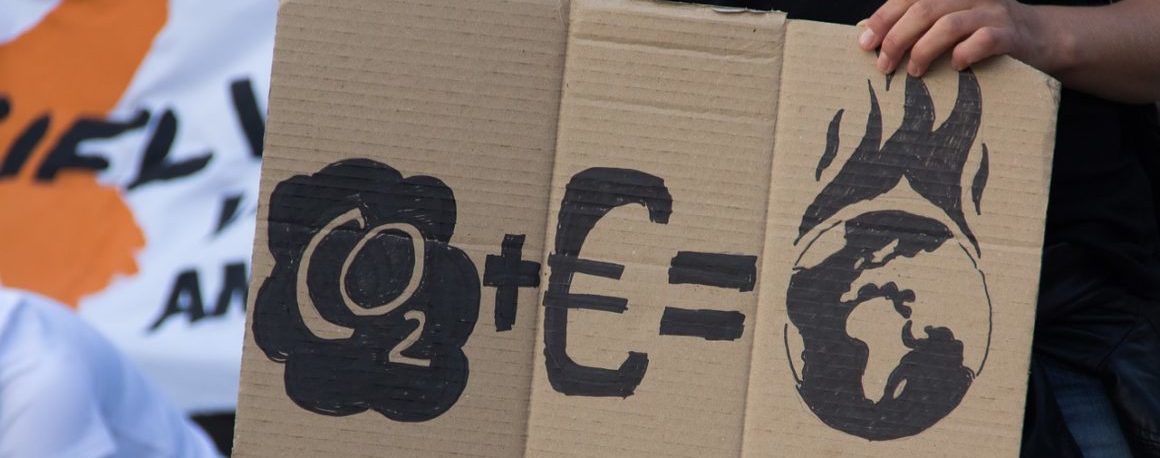 Close-up shot of a hand-drawn cardboard sign held by a person wearing jeans. The sign has symbols for CO2, a money symbol and then an equal sign. The answer is a picture of the world on fire.