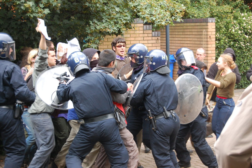 Large group of protestors surrounded by police officers in riot gear. The police have their backs to the camera and seem to be pushing on the protestors. One of the protestors is yelling.