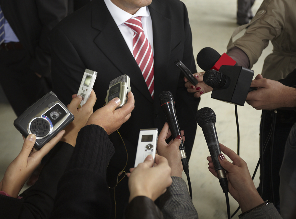 photograph of reporters' recording devices pushing for response from suited figure