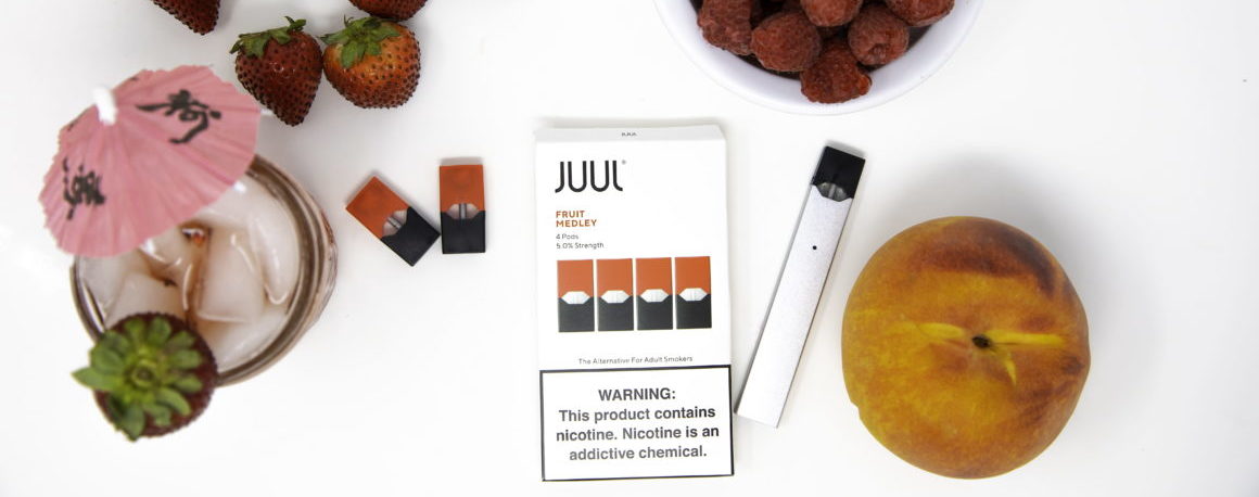 A view from above looking onto a white surface with berries, a peach, an alcoholic drink and an e-cigarette artfully arranged.