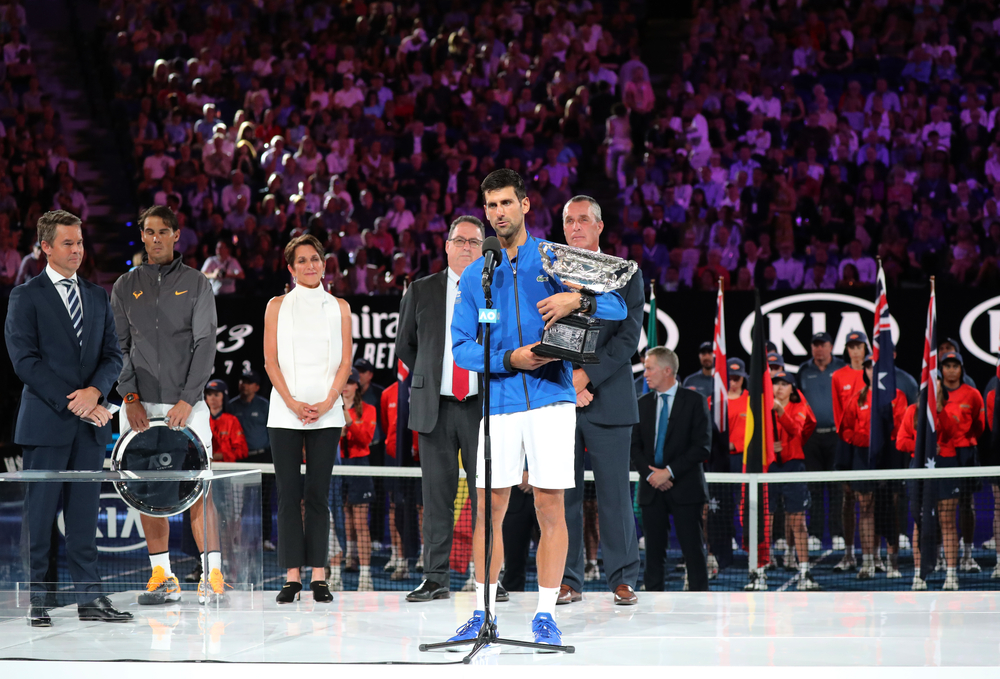 photograph of Djokovic on stage at mic with trophy in front of packed stadium