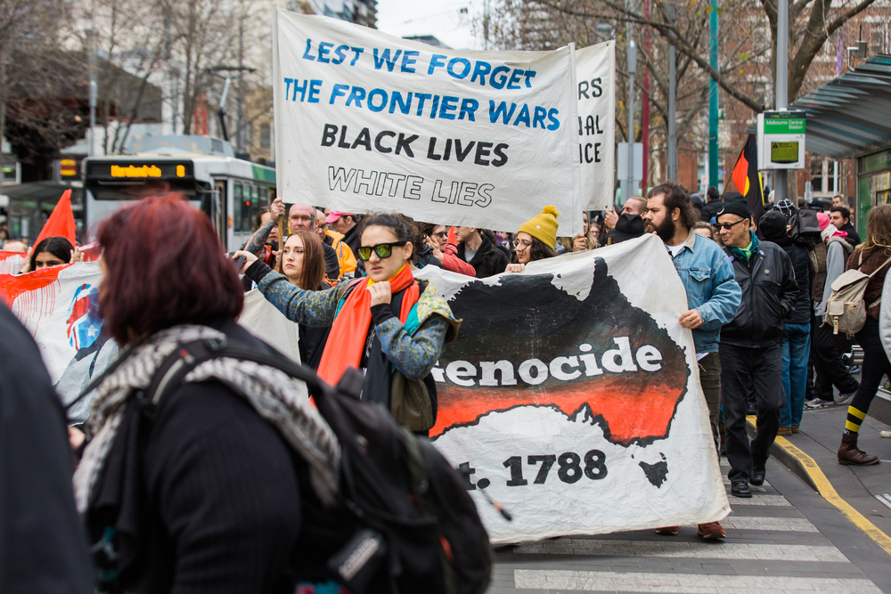 Protest in Australia; two signs are visible: one reads "lest we forget the frontier wars, black lives, white lies" and one shows a black and red image of Australia with the word "genocide" written on it