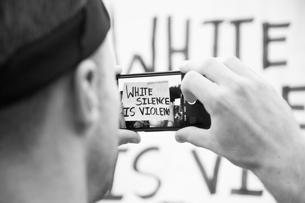 black-and-white photograph of protestor taking photo of "White Silence is Violence" sign with phone